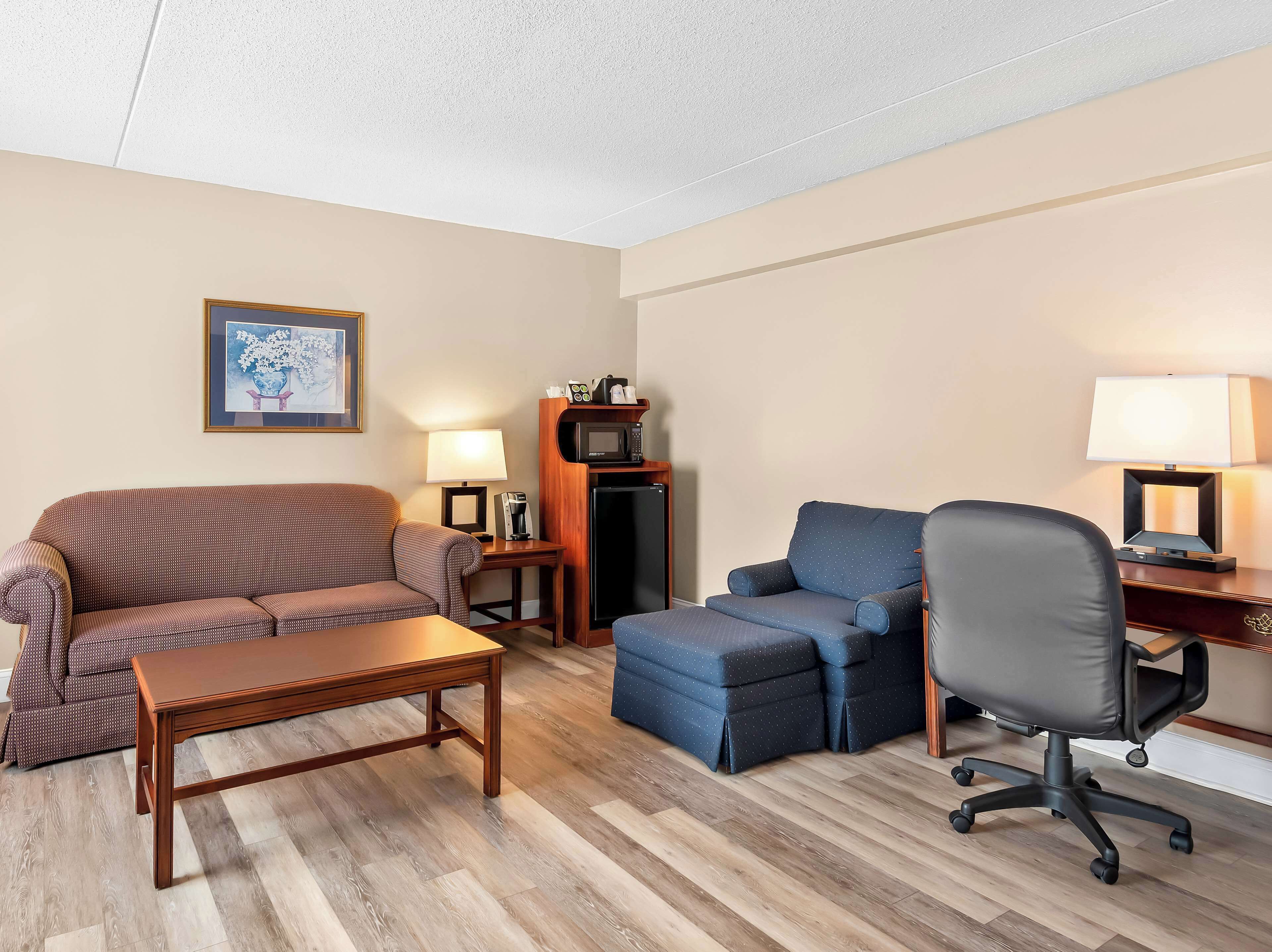 A hotel room with a sofa, a small table, a desk with a chair, a microwave, a refrigerator, and a lamp.