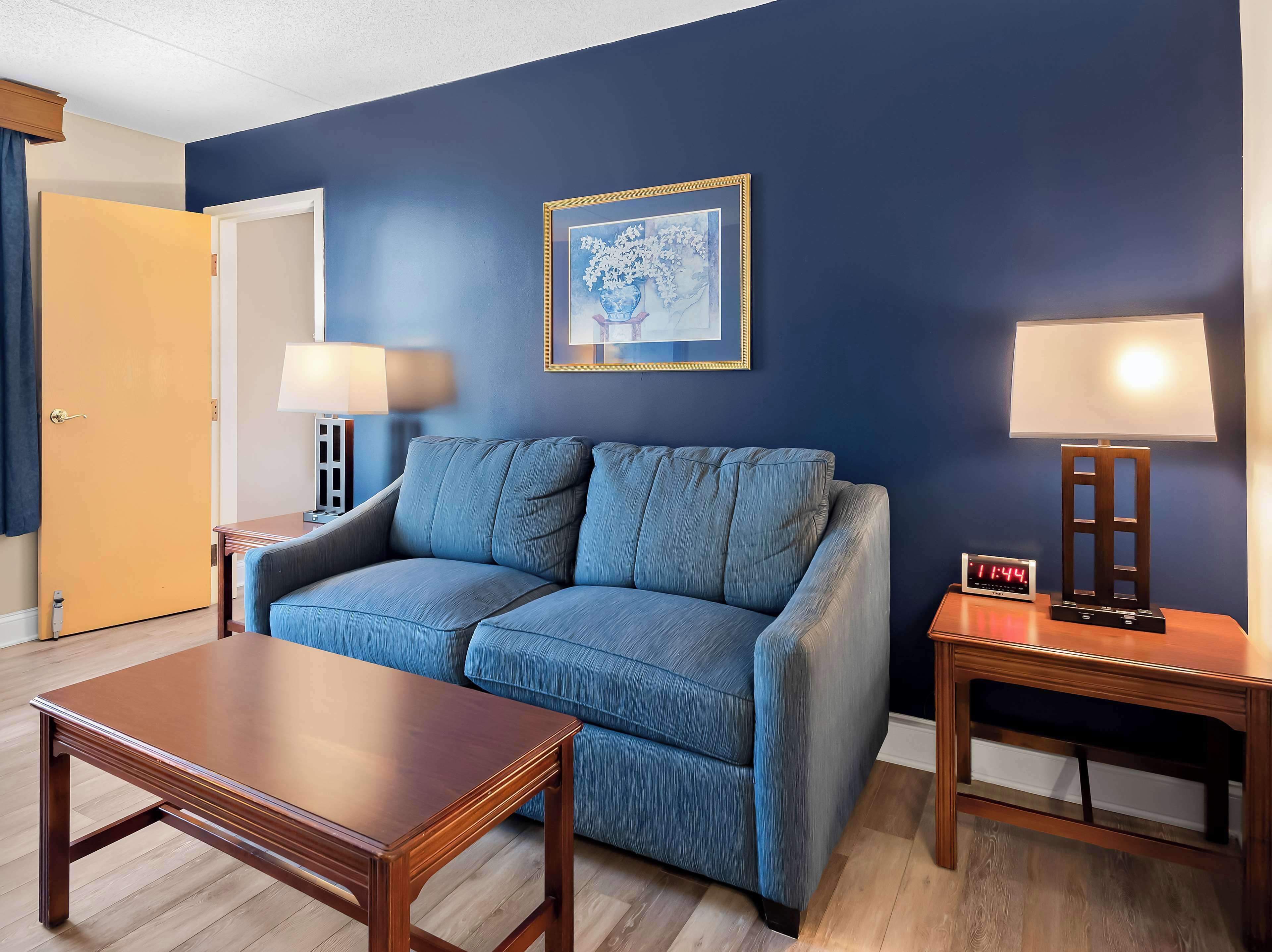 A room with a blue sofa, a wooden coffee table, two bedside tables with lamps, a framed picture on the wall, and a digital clock.