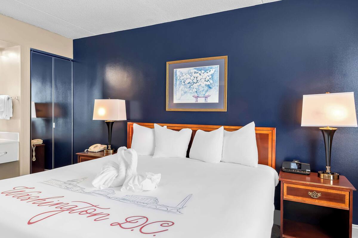 A hotel room with a bed, two bedside tables, a nightstand, a framed artwork, and a wall-mounted telephone.