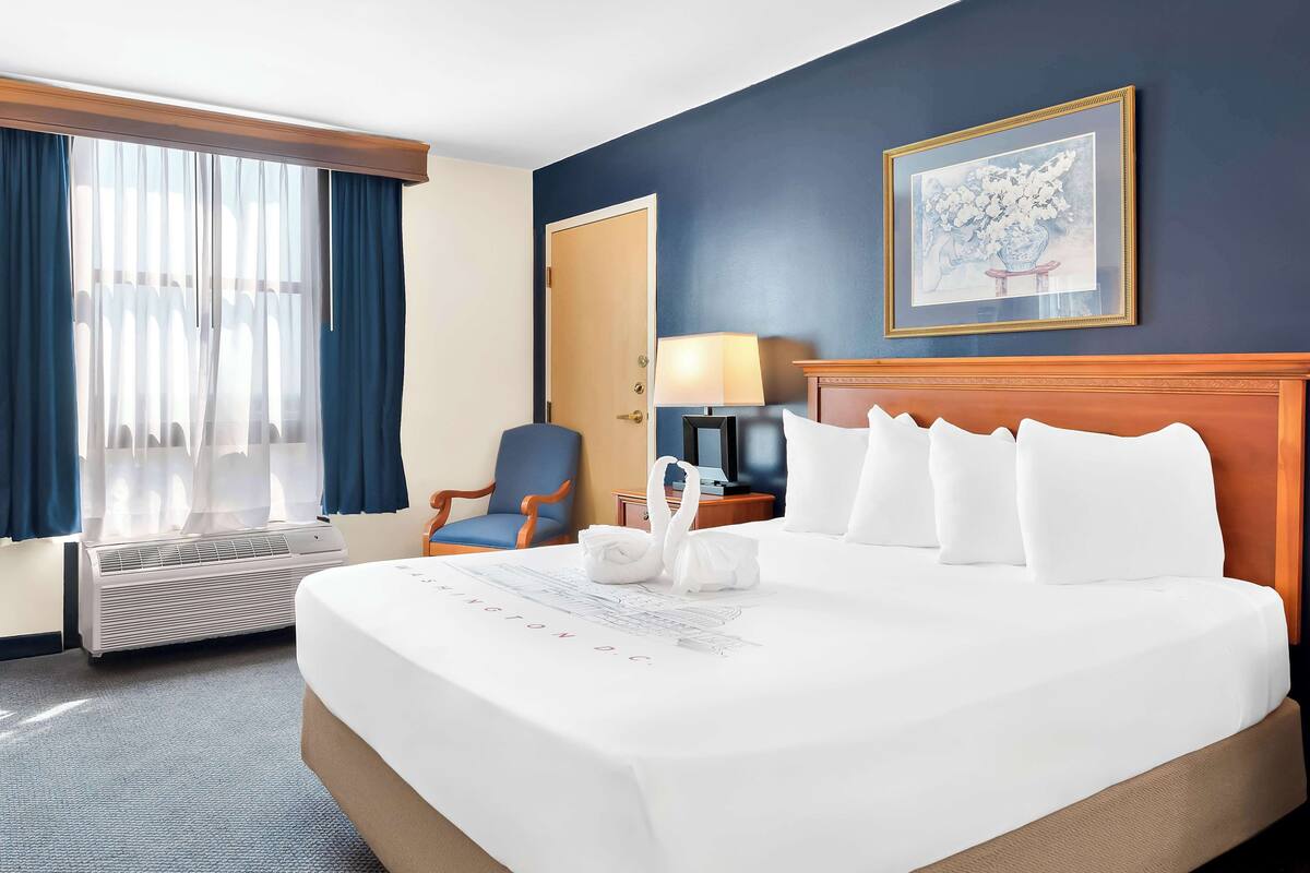 A hotel room with a large bed, a chair, a desk with a lamp, and a framed picture on the wall.