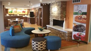 A modern living area with a blue sofa, a patterned ottoman, and a small round table. A kitchen is visible in the background with a stone wall and a fireplace. - Quality Inn Cranberry Township (Cranberry Township)