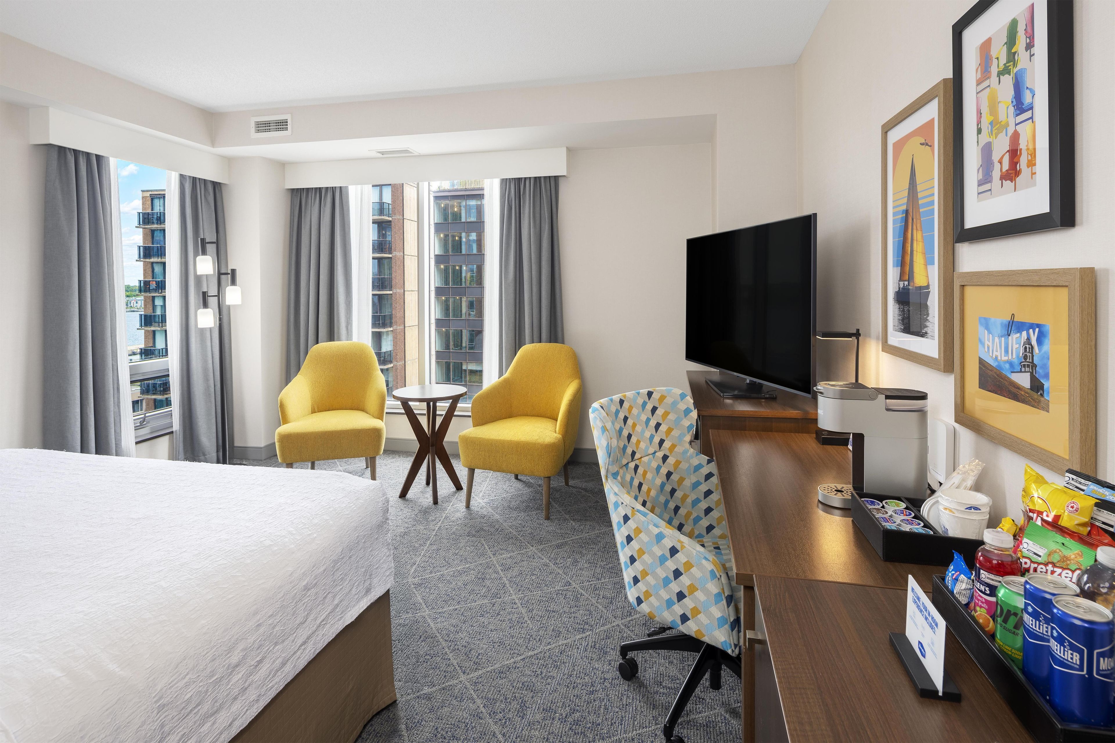 A hotel room with a bed, two yellow armchairs, a TV, and a minibar.