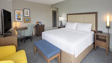 A hotel room with a large bed, a desk with a TV, a chair, a bedside table with a lamp, and a small table with a sign.