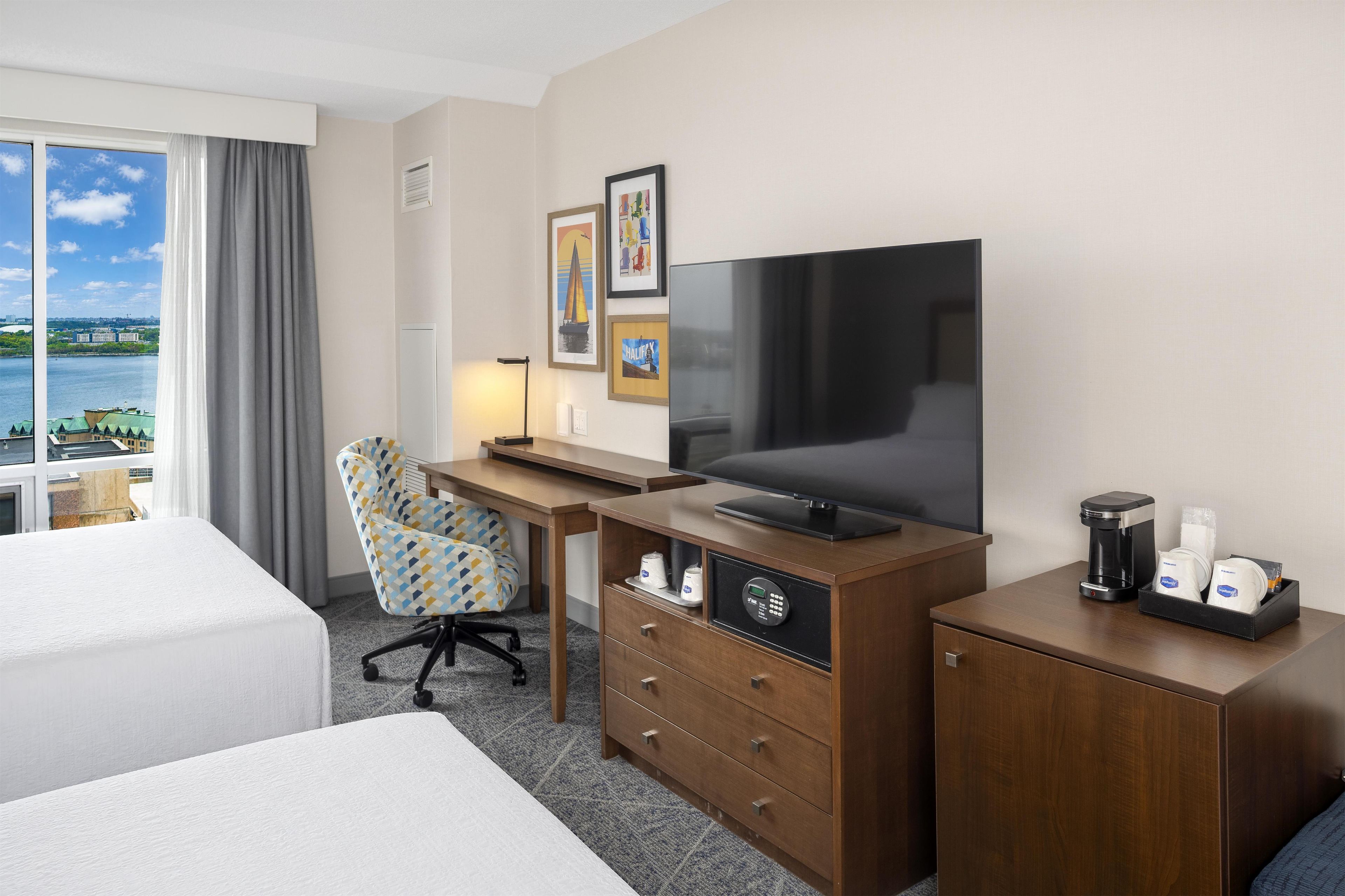 A hotel room with a bed, a desk with a television, a chair, a coffee maker, and a view of a body of water.