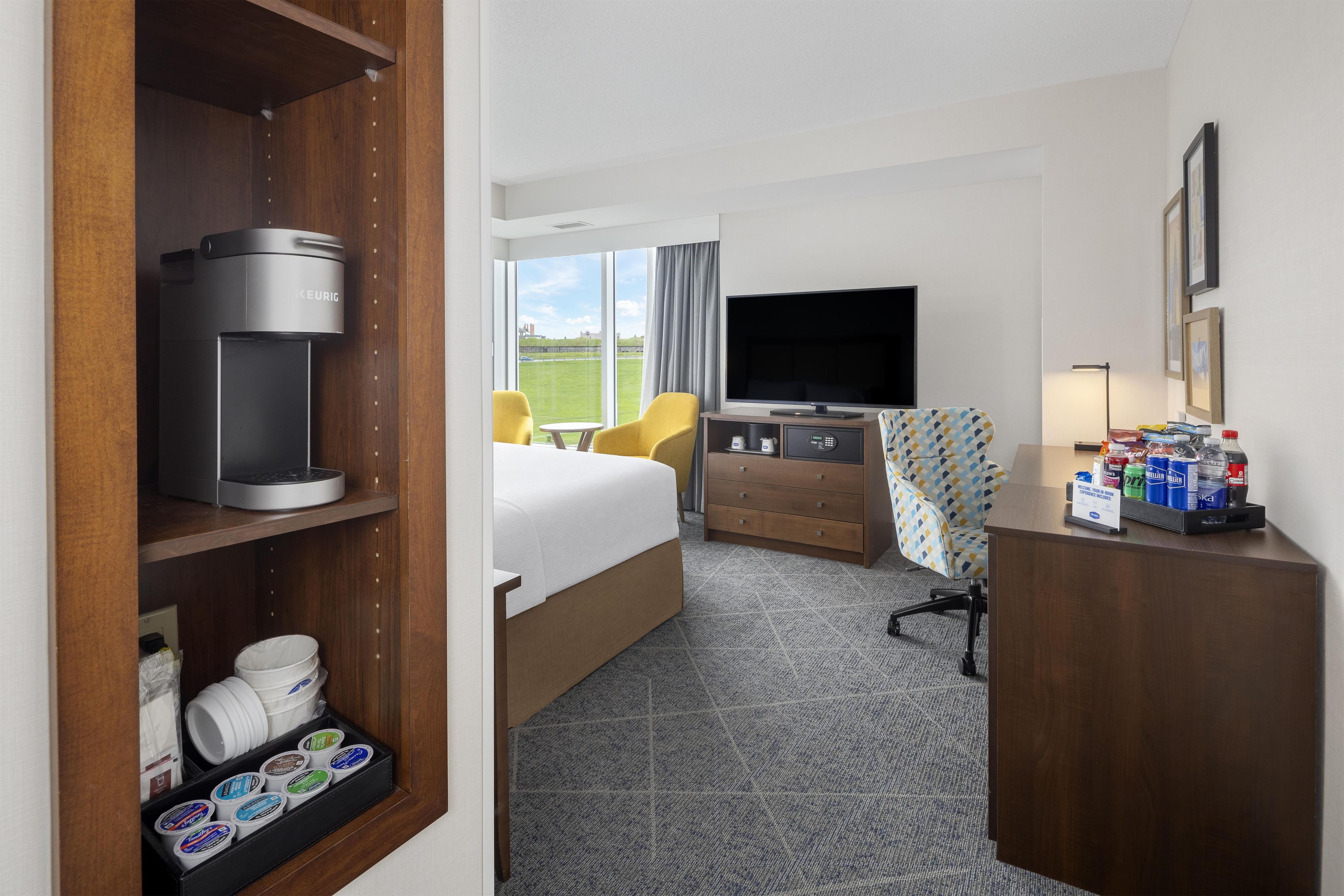 A hotel room with a bed, a desk with a flat-screen TV, a coffee maker, a minibar, and a view of the outdoors.