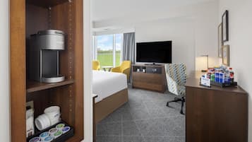 A hotel room with a bed, a desk with a flat-screen TV, a coffee maker, a minibar, and a view of the outdoors.