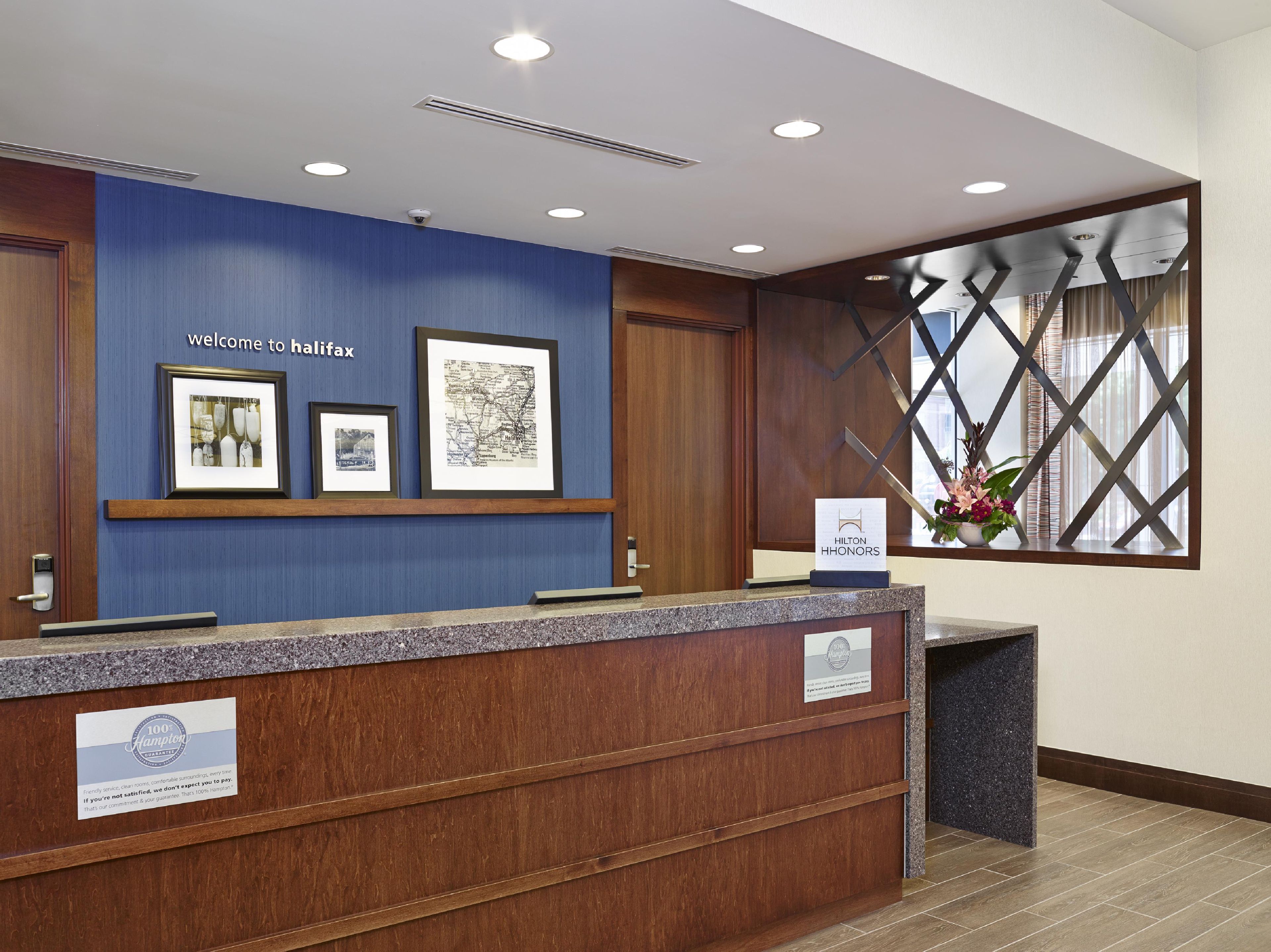 A hotel reception desk with framed pictures on the wall, a 'Welcome to Halifax' sign, and a 'Hilton Honors' sign.