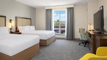 Quarto de hotel com duas camas, uma escrivaninha, uma cadeira, televisão e janela com vista para edifícios.