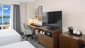 A hotel room with a bed, a desk with a television, a chair, a coffee maker, and a view of a body of water.