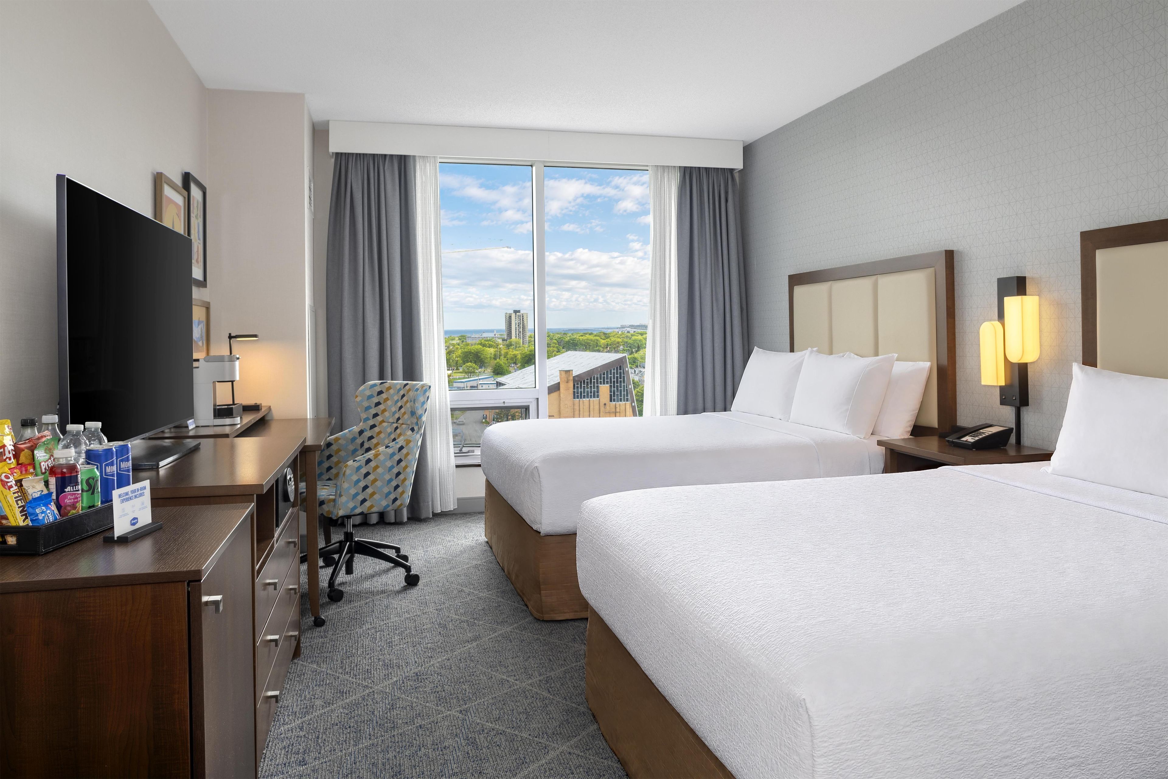 A hotel room with two beds, a desk with a flat-screen TV, a chair, and a view of the cityscape.
