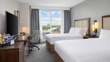 A hotel room with two beds, a desk with a flat-screen TV, a chair, and a view of the cityscape.