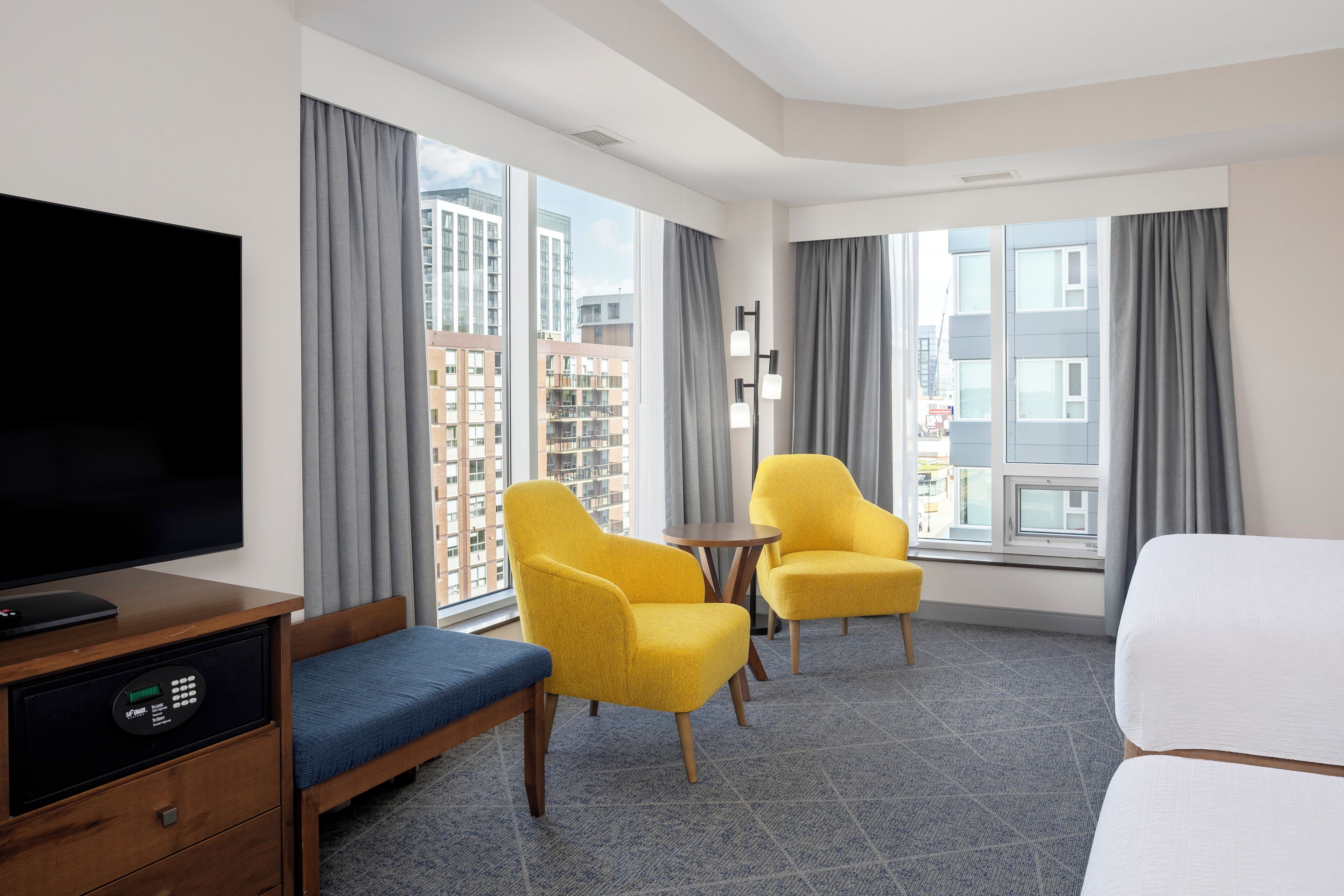 Habitación de hotel con una cama, un televisor, dos sillones amarillos, una mesita y ventanas amplias con cortinas.