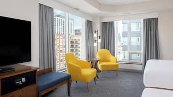 A hotel room with a bed, a TV, two yellow armchairs, a small table, and large windows with curtains.