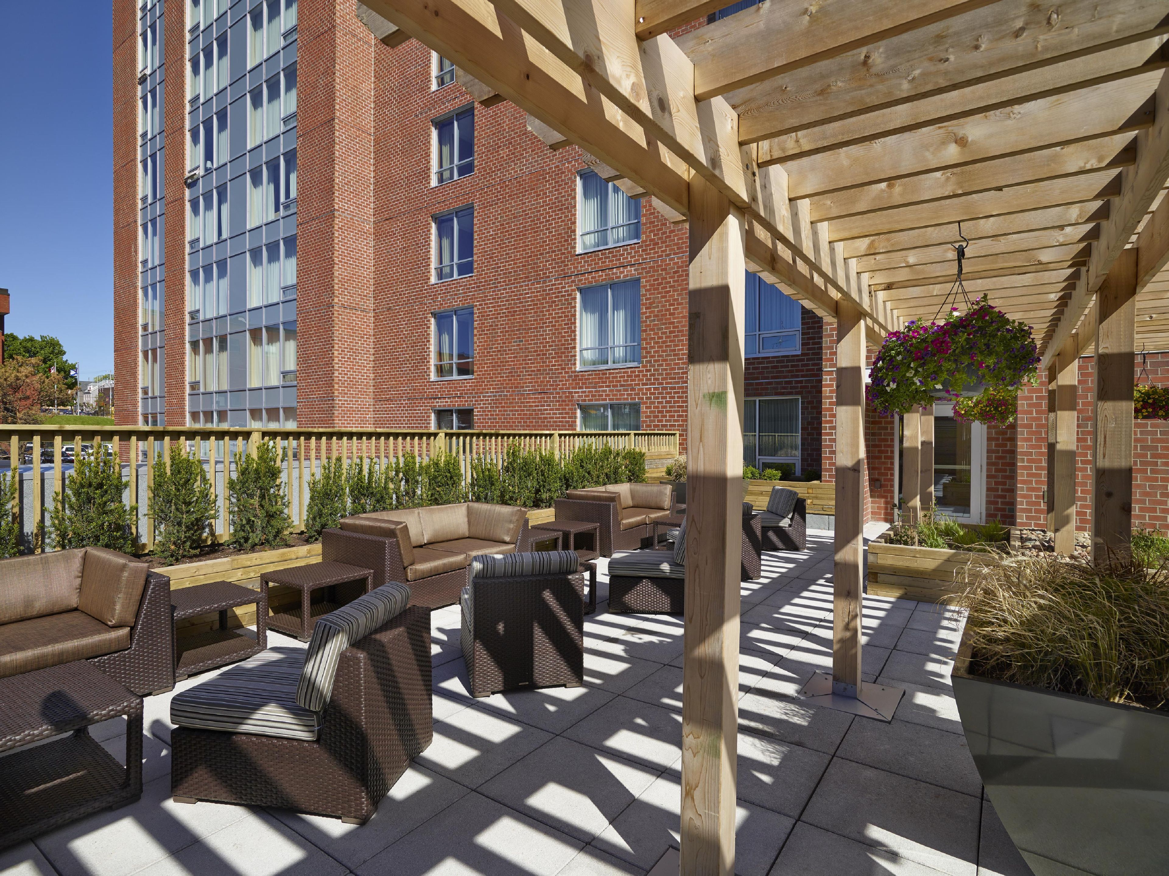 An outdoor patio area with wicker furniture, a wooden pergola, and a brick building in the background.