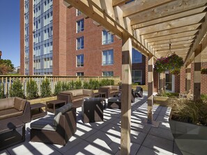 An outdoor patio area with wicker furniture, a wooden pergola, and a brick building in the background.