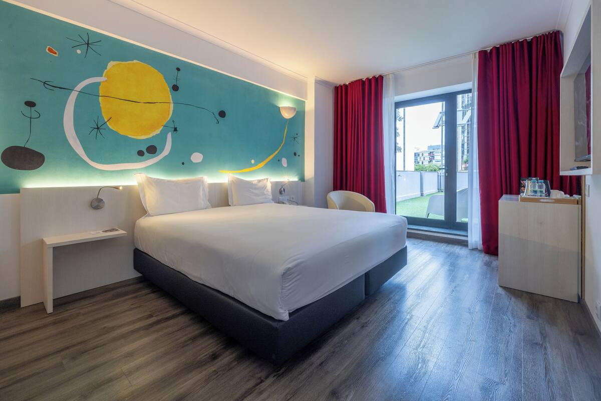 Ein modernes Hotelzimmer mit einem großen Bett, einem Schreibtisch, einem Wandbild und einem Balkon mit roten Vorhängen.