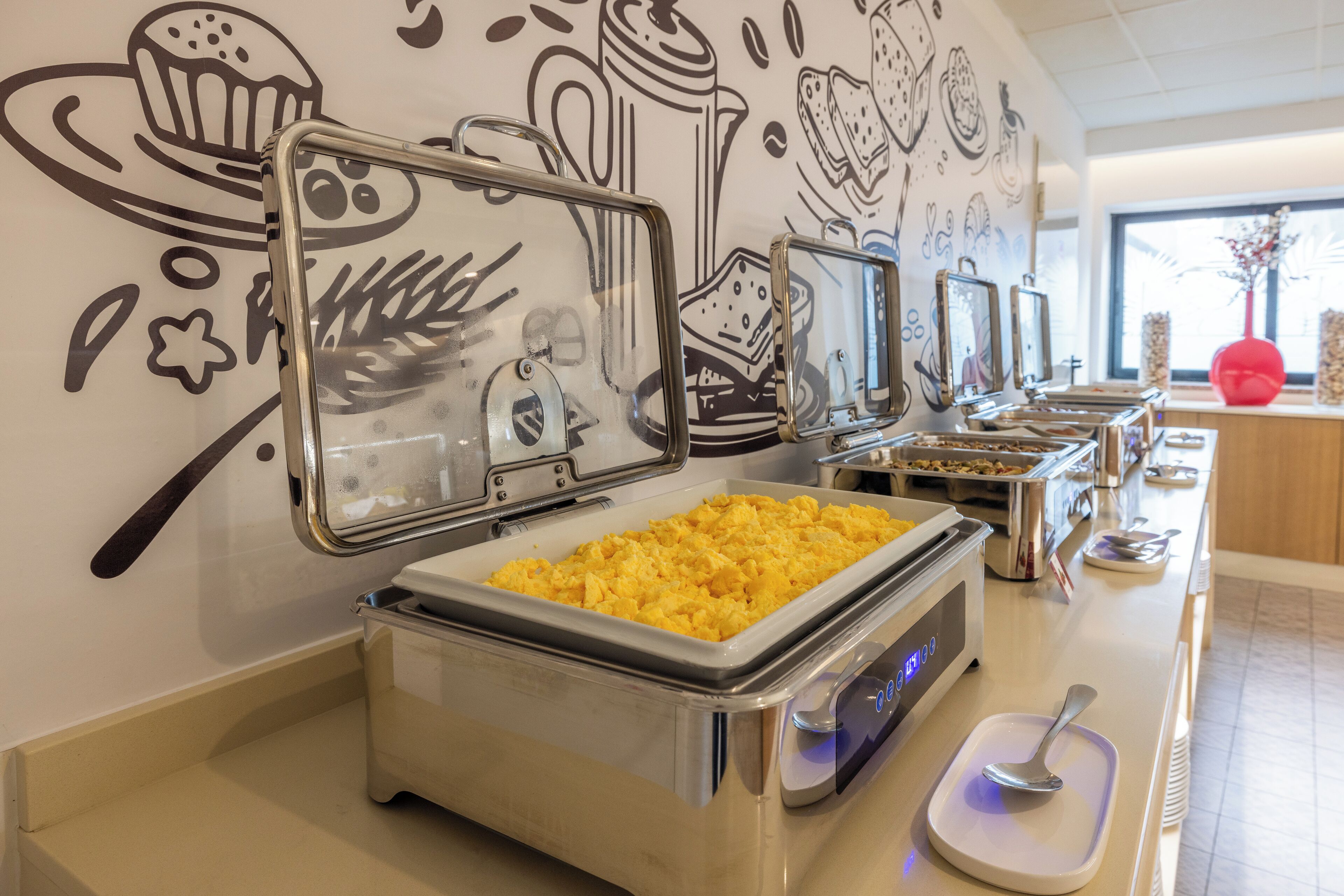 A buffet area with various food options, including scrambled eggs, in a well-lit room with a wall decorated with food illustrations.