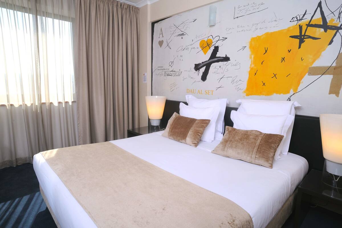 Ein Hotelzimmer mit einem großen Bett, beiger Bettwäsche, einer Wand mit Kunstwerken und Unterschriften sowie einem Fenster mit Vorhängen.