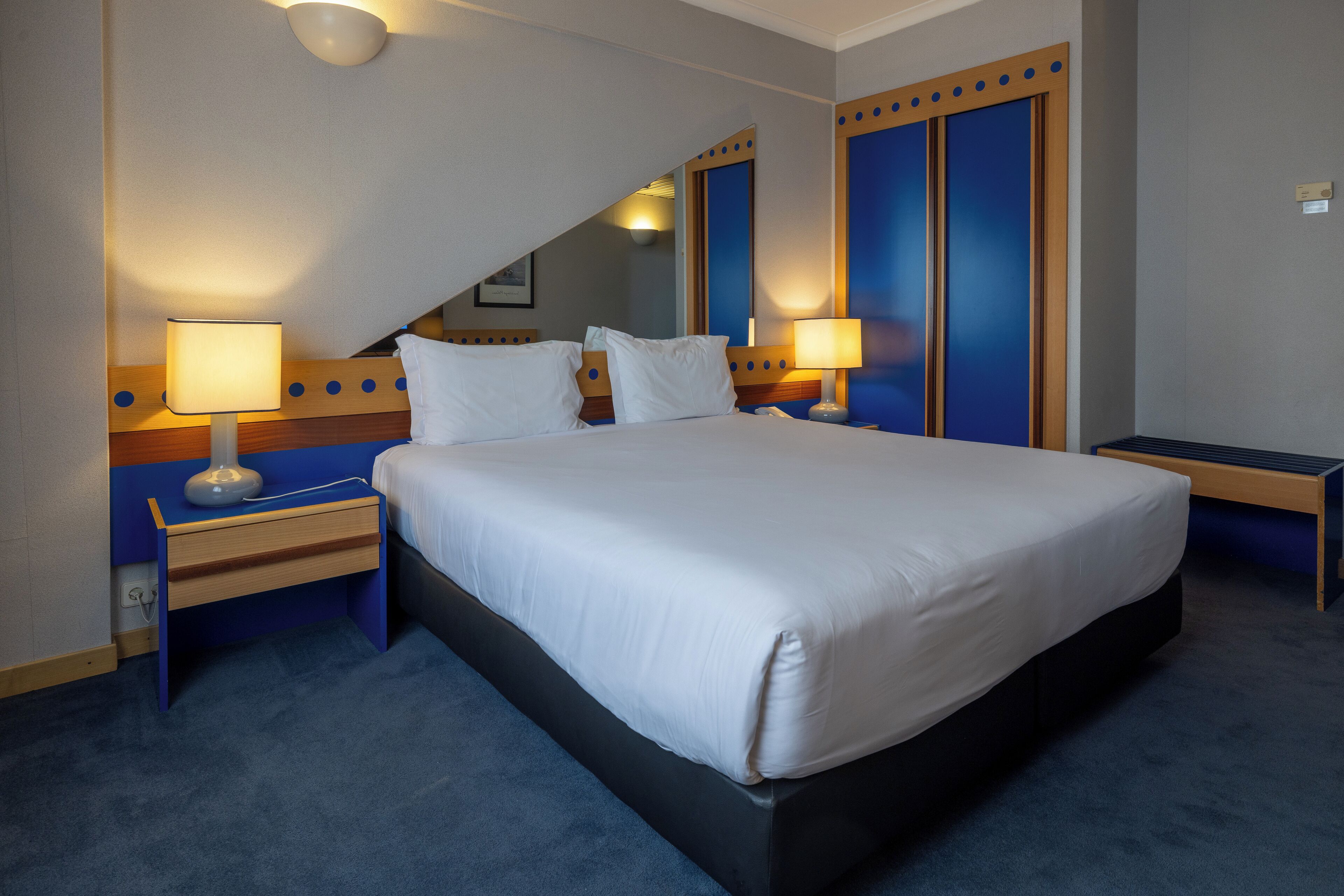 Une chambre d’hôtel avec un grand lit, deux tables de chevet avec des lampes, une tête de lit en bois et un mur d’accent bleu.