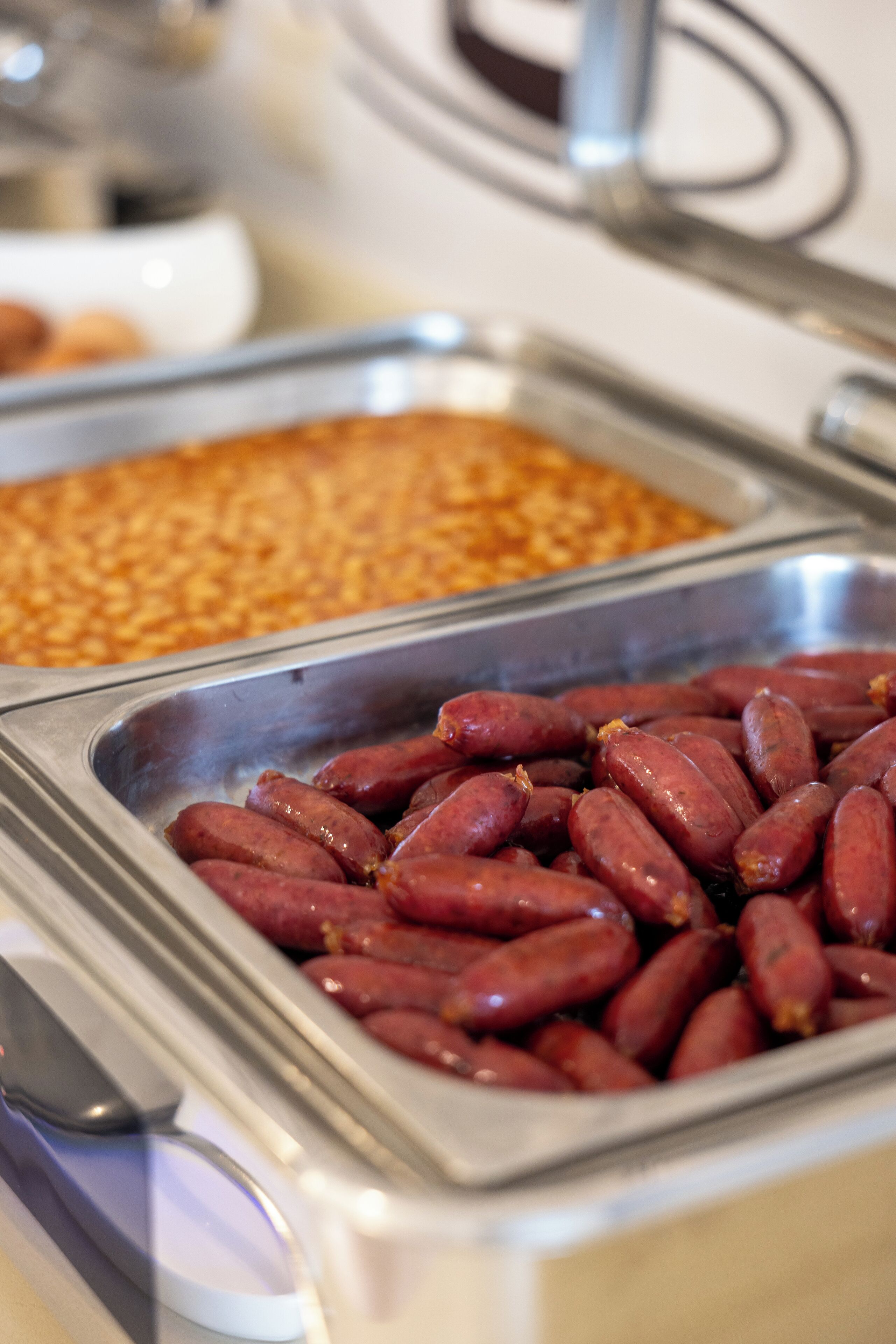 A buffet with various food items, including a tray of red sausages and a container of what appears to be a grain dish.