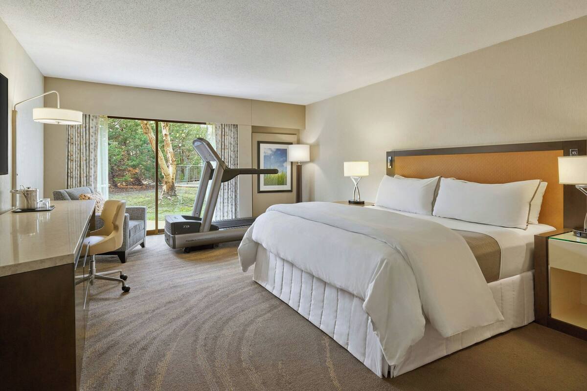 Ein Hotelzimmer mit einem großen Bett, einem Schreibtisch, einem Sessel, einem Laufband und einem großen Fenster mit Blick auf Bäume.