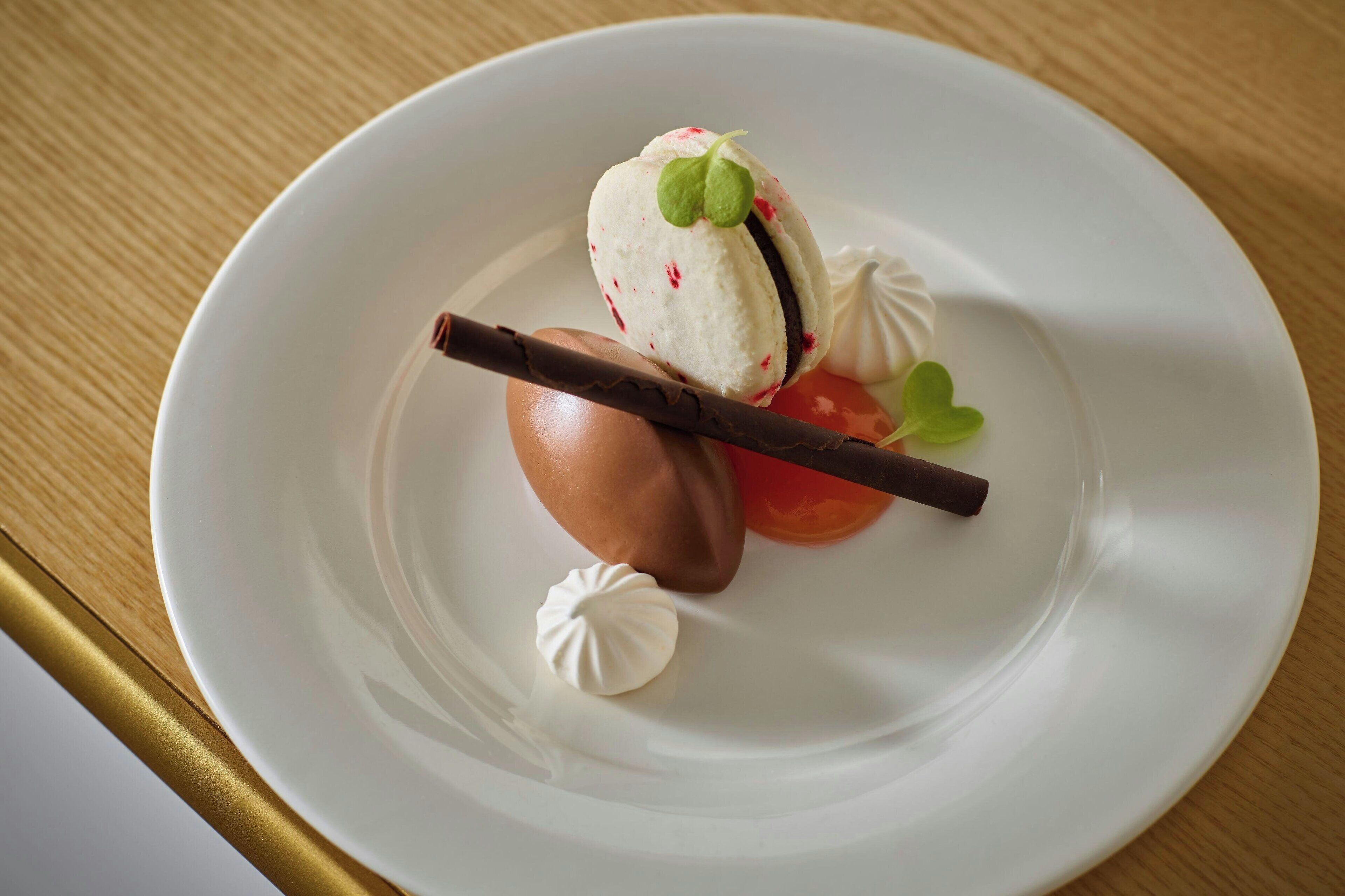 A dessert on a white plate with a chocolate piece, a small green leaf, and a dollop of cream.
