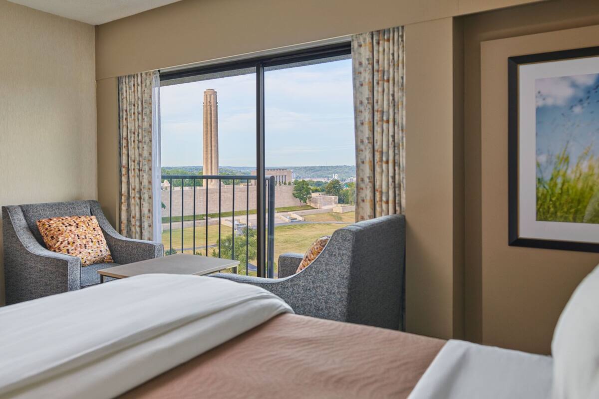 Ein Hotelzimmer mit einem großen Bett, zwei Sesseln, einem kleinen Tisch und Blick auf ein weit entferntes Gebäude durch ein großes Fenster.