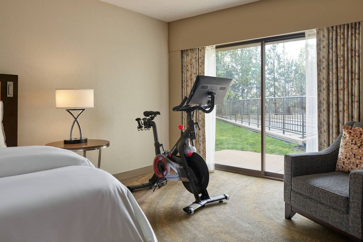 Ein Hotelzimmer mit Bett, Schreibtisch mit Lampe, Heimtrainer, grauem Sessel und großem Fenster mit Blick auf einen Balkon und Grünflächen.