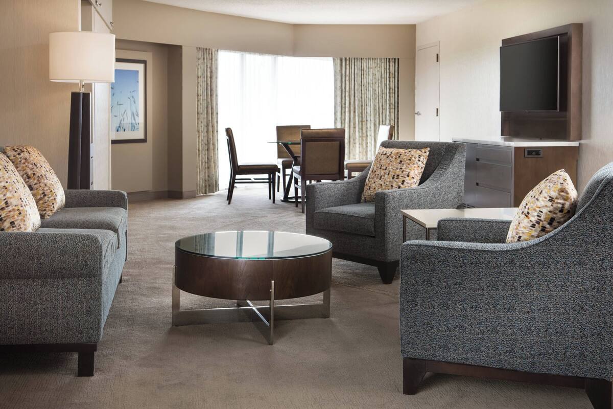 Ein modernes Hotelzimmer mit einer Sitzecke, ausgestattet mit zwei Sofas, einem Glastisch und einem Esstisch mit Stühlen. Ein Flachbildfernseher ist an der Wand befestigt, und es gibt ein großes Fenster mit Vorhängen.