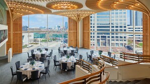 A spacious dining area with multiple tables set for guests, large windows offering a city view, and a distinctive wooden ceiling design. - The Westin Kansas City at Crown Center (Kansas City)