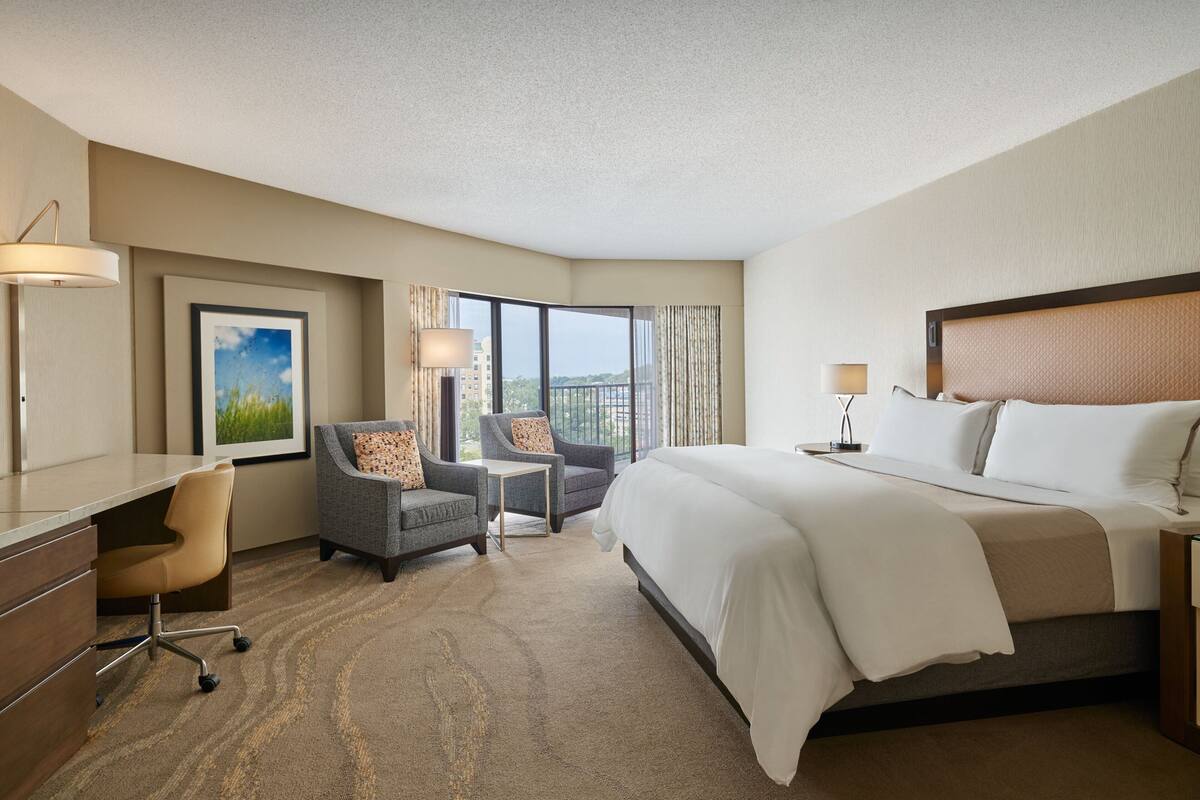 Ein Hotelzimmer mit einem großen Bett, zwei Sesseln, einem Schreibtisch und Blick auf die Stadt durch große Fenster.