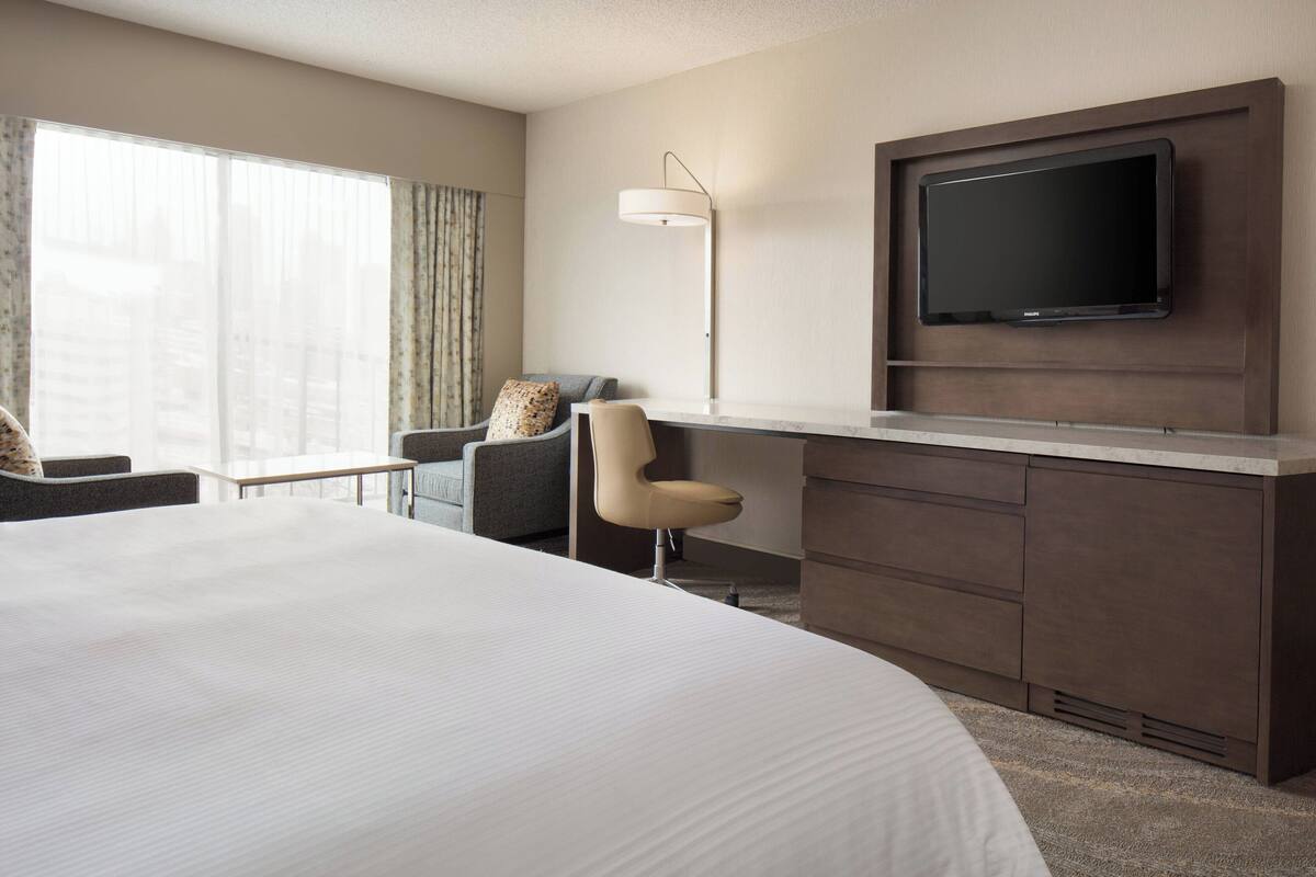 Ein Hotelzimmer mit einem großen Bett, einem Schreibtisch, einem Flachbildfernseher, einem Sessel und einer Lampe.