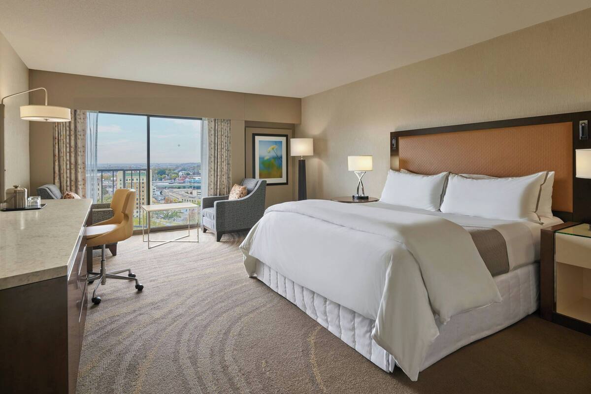Ein Hotelzimmer mit einem großen Bett, einem Schreibtisch, einem Stuhl und Blick auf die Stadt durch das Fenster.