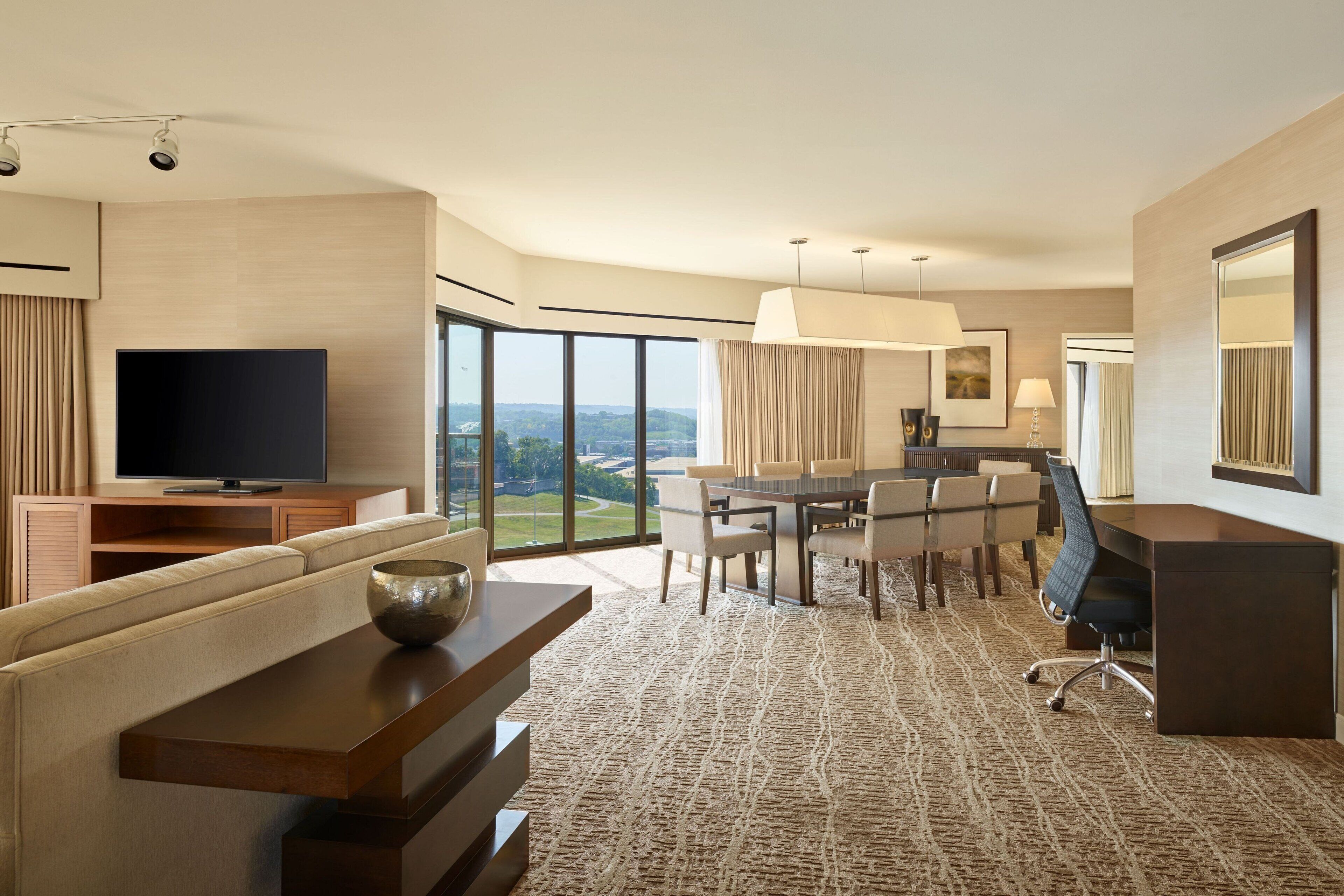 A hotel room with a large flat-screen TV, a dining area with a table and chairs, a sofa, and a large window offering a scenic view.
