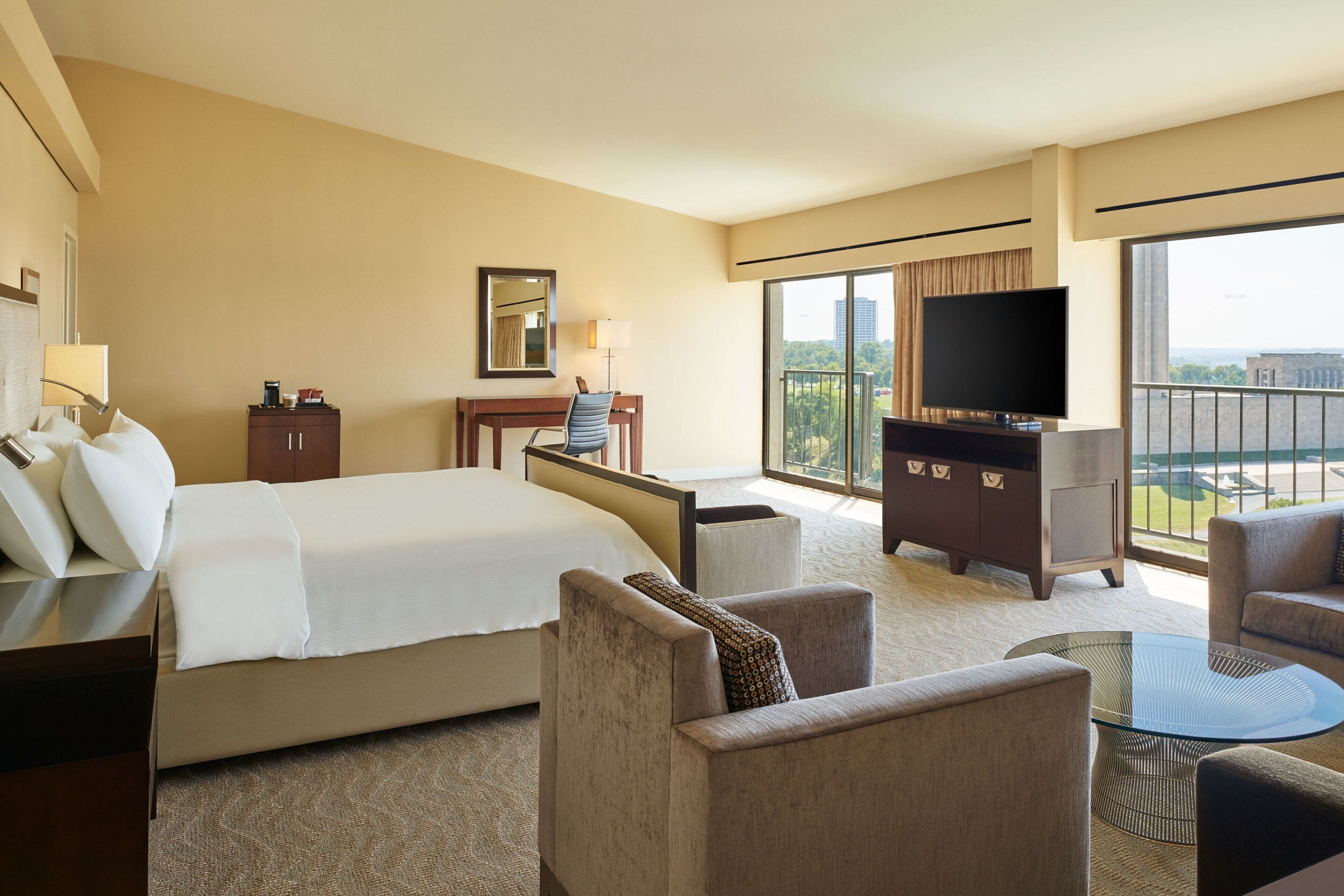 A hotel room with a large bed, a desk with a chair, a television, and a balcony with a view.