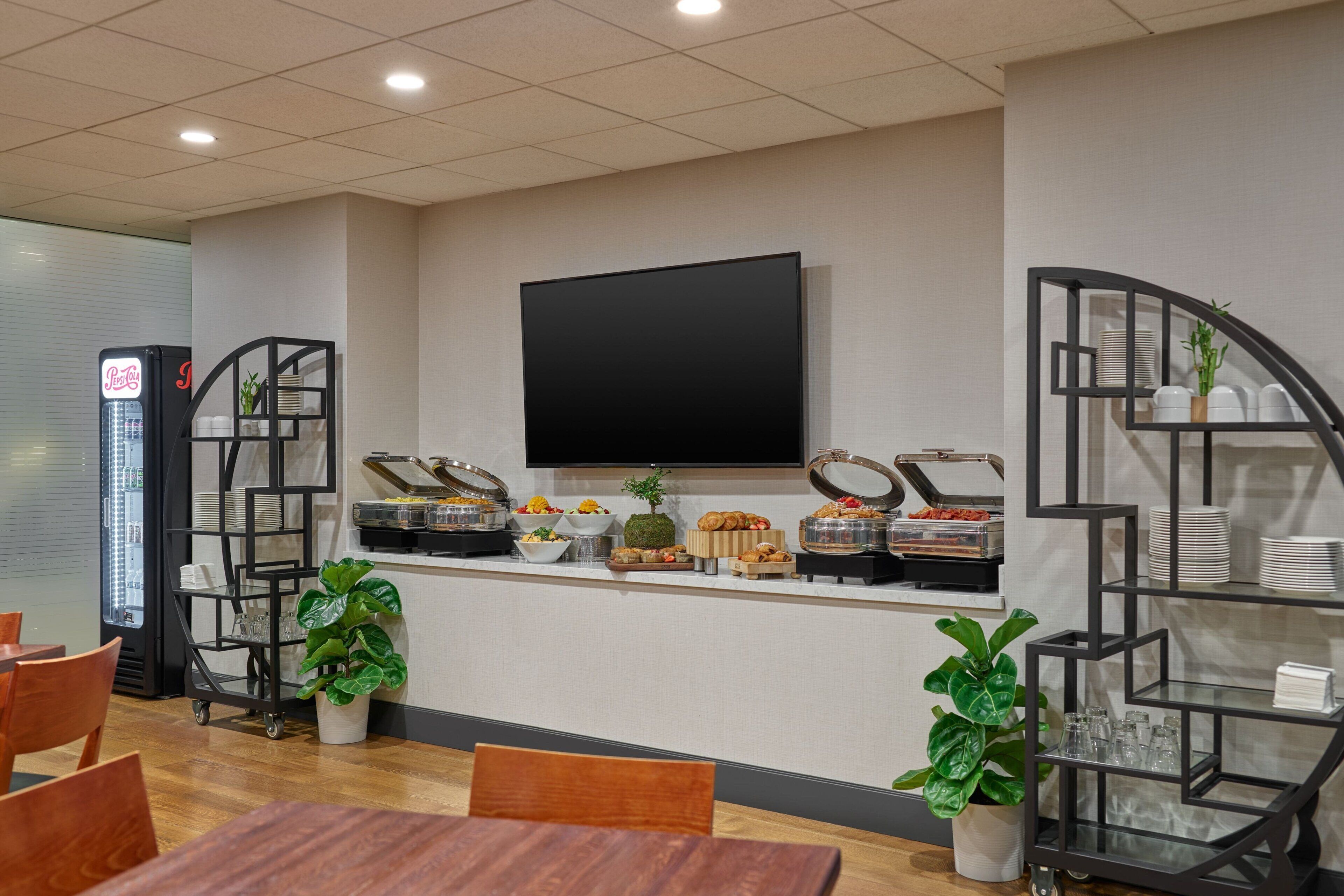A modern hotel room with a flat-screen TV mounted on the wall, a buffet counter with various food items, and a seating area with wooden tables and chairs.