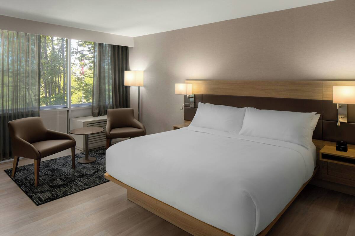 A modern hotel room with a large bed, two armchairs, a small table, and a view of trees through the window.
