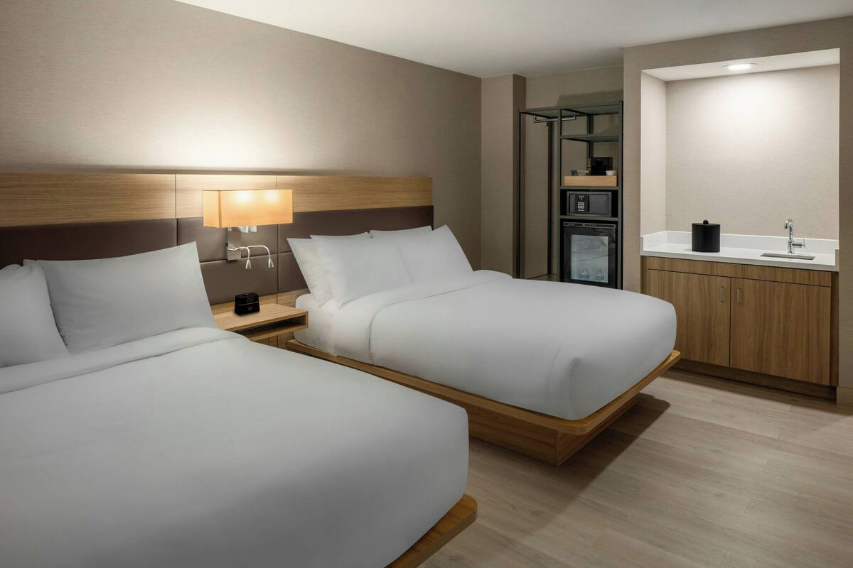 A hotel room with two beds, a bedside table with a lamp, a small kitchenette, and a bathroom with a sink and mirror.