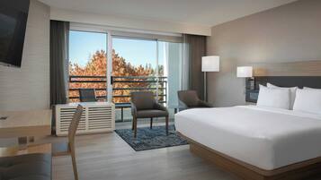 A modern hotel room with a large bed, a desk, a chair, and a view of autumn trees through a balcony.