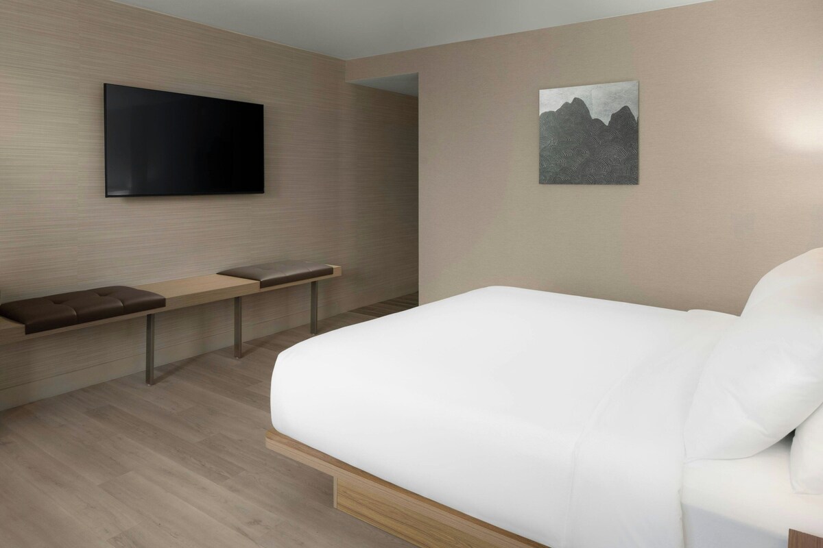 A modern hotel room with a large bed, a flat-screen TV mounted on the wall, a bench with cushions, and a painting on the wall.