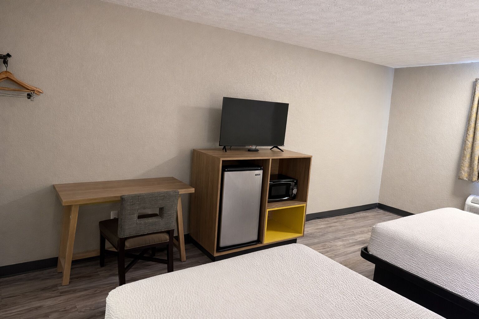 A hotel room with a wooden desk, a television on a shelf, a refrigerator, a microwave, a chair, and two beds.