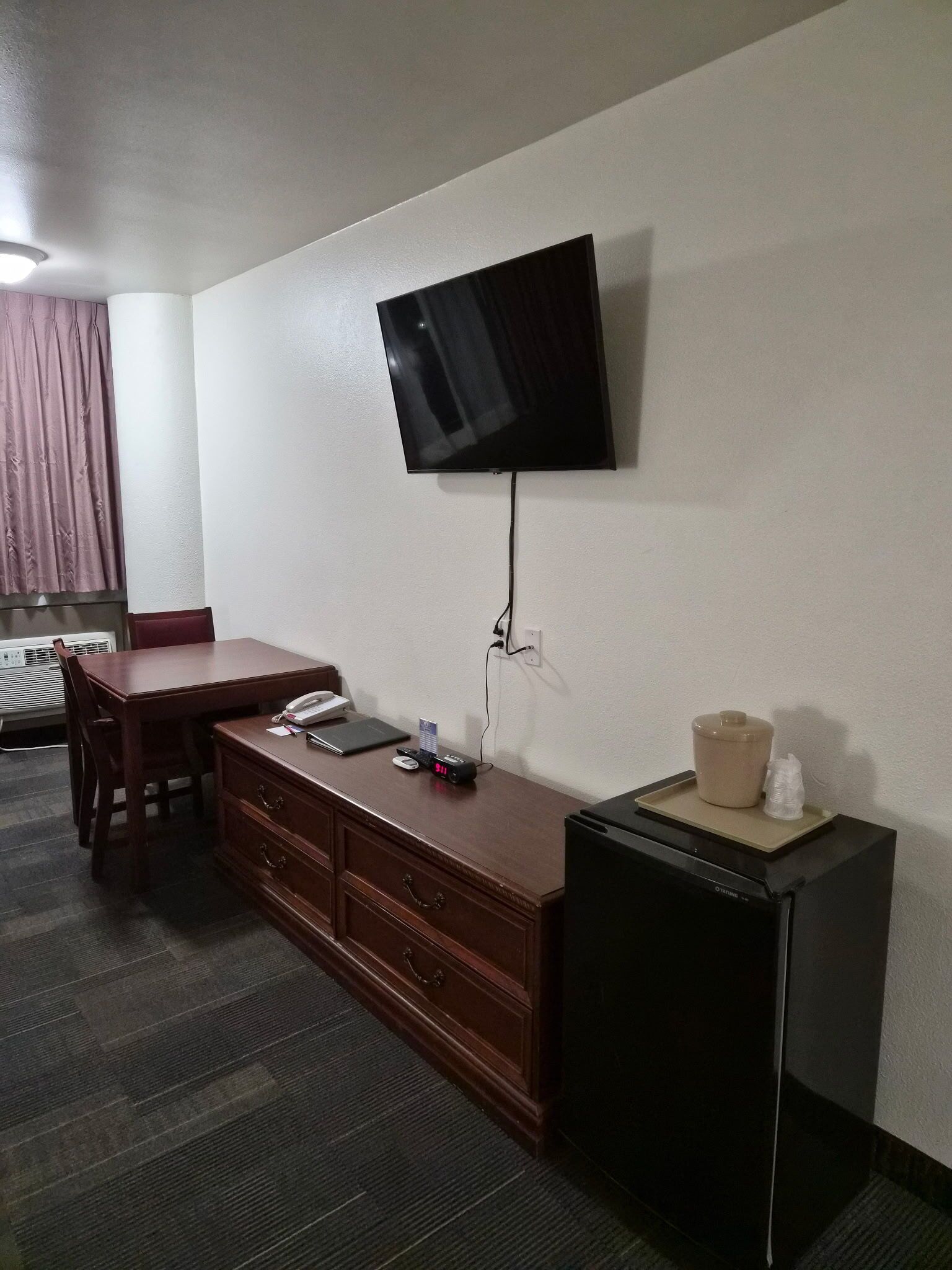 Quarto de hotel com televisão de tela plana na parede, escrivaninha de madeira com telefone, pequeno frigorífico e micro-ondas.
