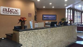 Un comptoir de réception d’hôtel avec une enseigne indiquant « Alex Hotel & Suites », un tableau encadré accroché au mur et un vase rempli de fleurs posé sur le comptoir.