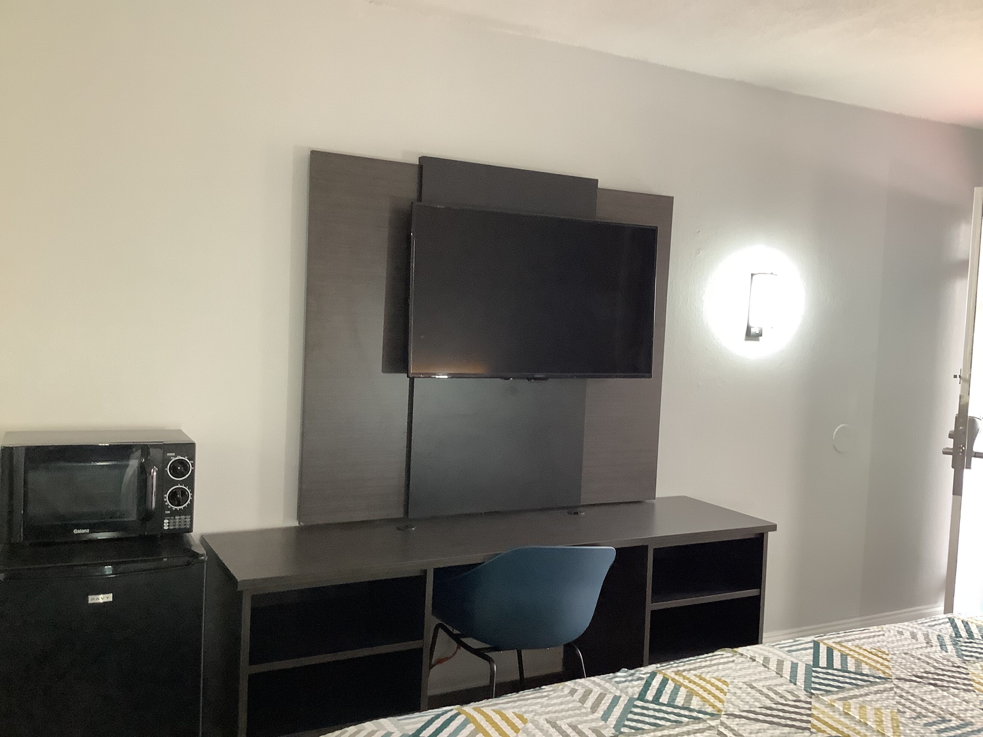 Habitación pequeña con cama, escritorio, silla, televisor montado en la pared y microondas sobre el escritorio.