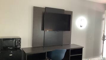 Habitación pequeña con cama, escritorio, silla, televisor montado en la pared y microondas sobre el escritorio.