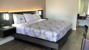A modern hotel room with a large bed, a nightstand, a bedside table, a built-in bench, a mirror, and a bathroom with a sink and toilet.