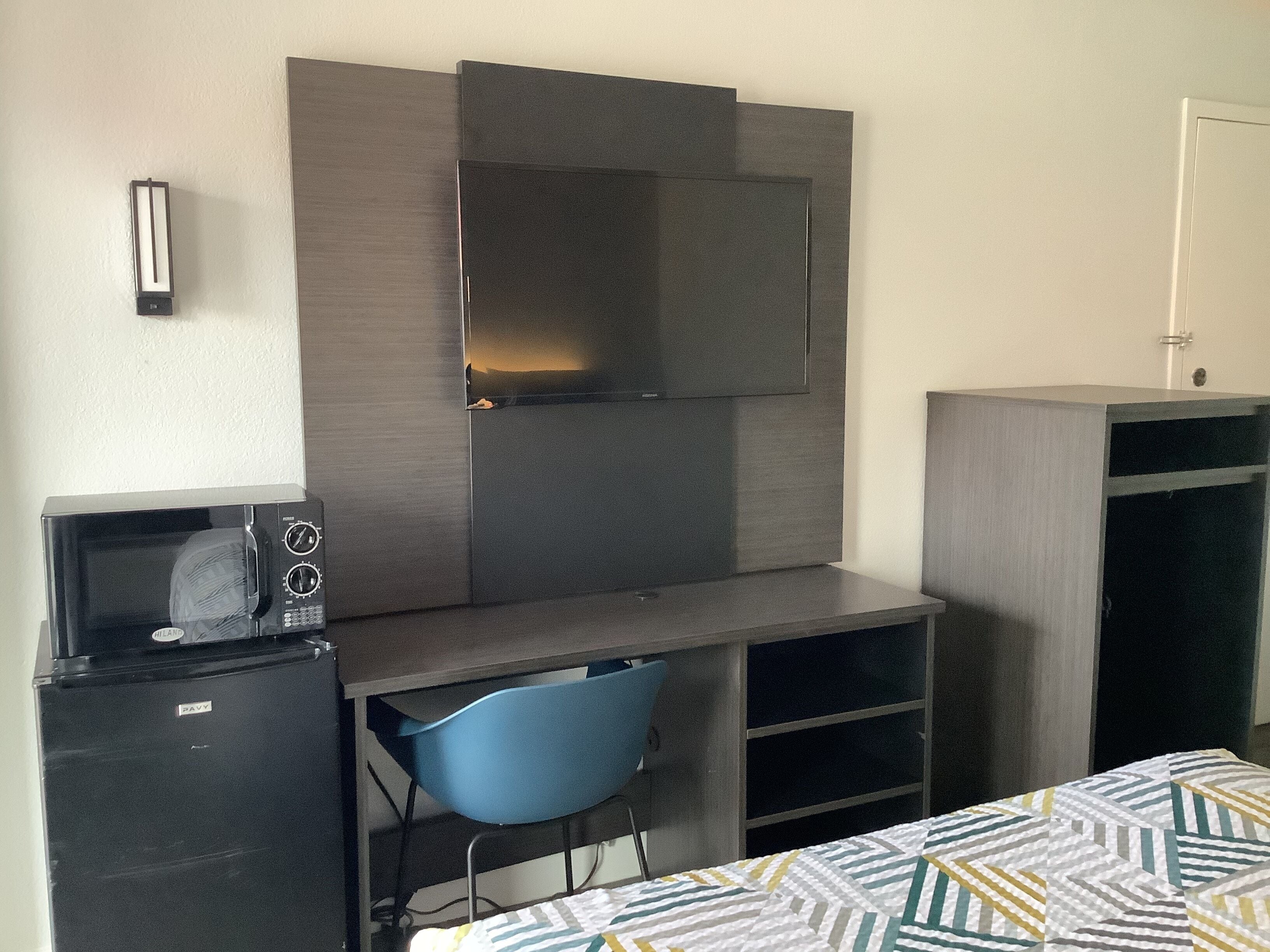 A compact hotel room with a kitchenette, a bed with a patterned duvet, a blue chair, a desk with a computer, and a TV mounted on the wall.