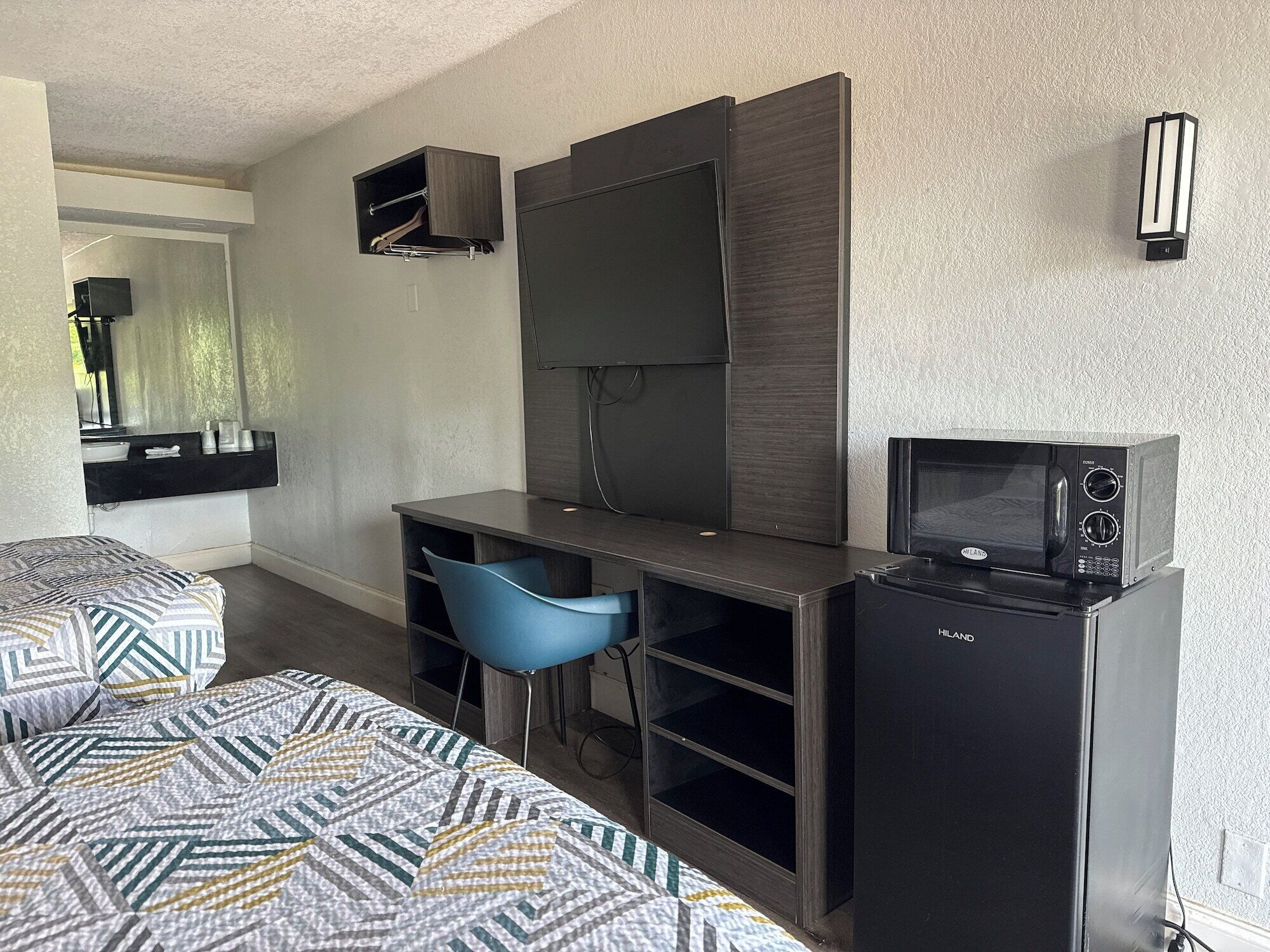 A compact hotel room with a bed, a desk with a chair, a flat-screen TV, a microwave, and a small shelf.