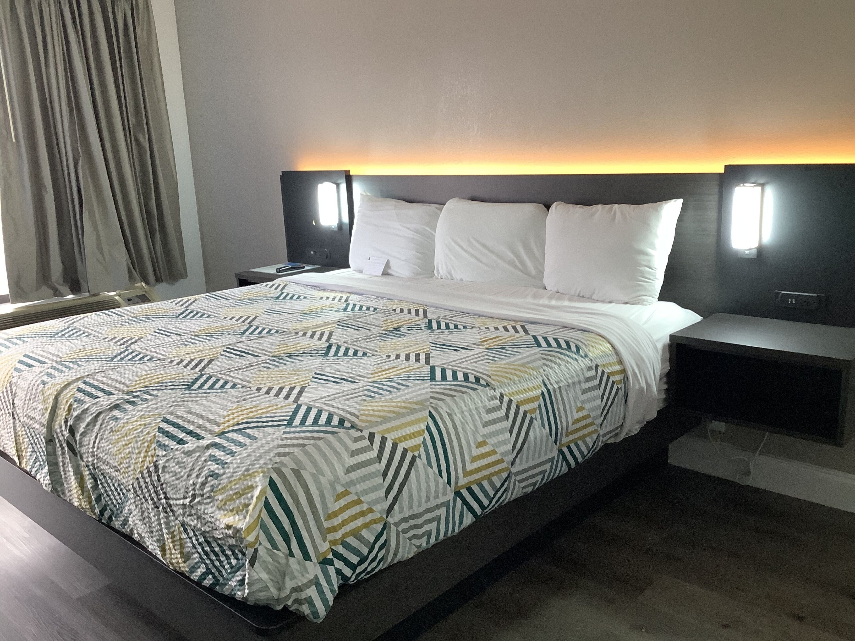Una habitación de hotel moderna con una cama grande, cabecero con iluminación integrada, mesita de noche y ventana con cortinas.