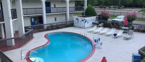 A swimming pool with lounge chairs and umbrellas, a covered area with a blue roof, and a fenced parking area.